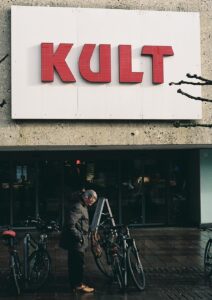 Ein großes Schild an einer Fassade mit der Aufschrift "Kult", davor ein Mann, der sein Fahrrad abstellt.